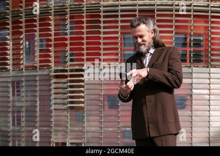 Gray haired bearded man in formal suit drinking takeaway coffee and browsing mobile phone on urban street Stock Photo