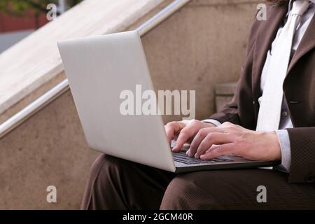 Cropped unrecognizable male entrepreneur in formal clothes sitting on stairway and working online on laptop in city Stock Photo