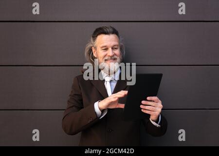 Elegant bearded mature man in classy formal suit standing near gray wall and browsing internet on digital tablet Stock Photo