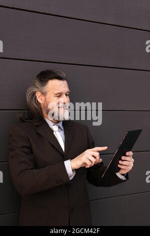 Elegant bearded mature man in classy formal suit standing near gray wall and browsing internet on digital tablet Stock Photo
