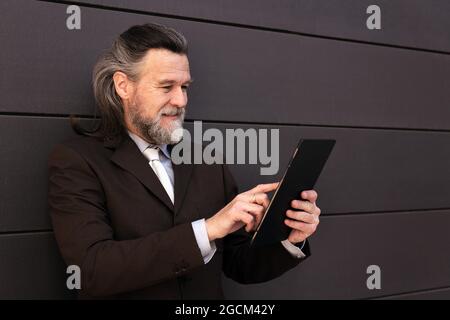 Elegant bearded mature man in classy formal suit standing near gray wall and browsing internet on digital tablet Stock Photo
