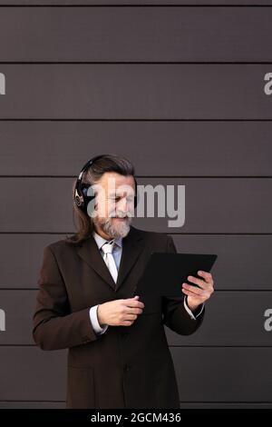 Content gray haired bearded male in formal suit and wireless headphones using tablet while communicating online against gray wall Stock Photo