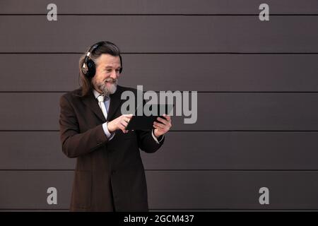 Content gray haired bearded male in formal suit and wireless headphones using tablet while communicating online against gray wall Stock Photo