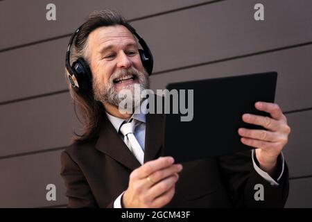 From below content gray haired bearded male in formal suit and wireless headphones using tablet while communicating online against gray wall Stock Photo