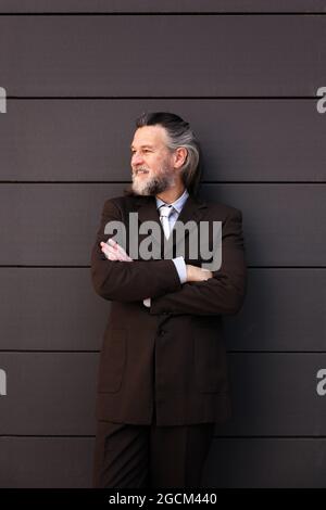 Confident successful mature bearded male in elegant suit standing with arms crossed and looking away with smile against gray wall Stock Photo