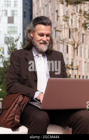 Happy successful gray haired bearded male in elegant suit using laptop while sitting on urban street Stock Photo