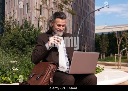 Happy successful gray haired bearded male in elegant suit drinking takeaway coffee using laptop while sitting on urban street Stock Photo