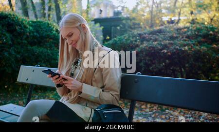 Young blonde girl uses a cell phone on a bench in the Park outdoors. Stock Photo