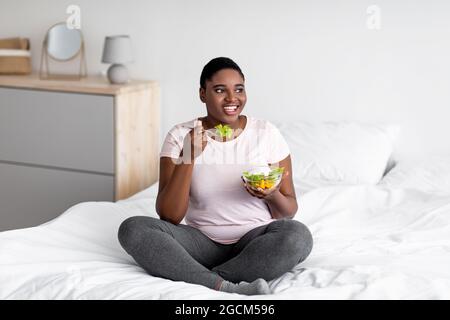 Balanced weight loss diet concept. Plus size Afro woman eating vegetable salad, sitting on bed at home Stock Photo