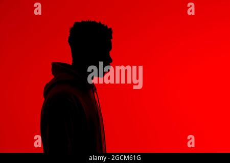 Side view of silhouette of unrecognizable African American male in hoodie standing on red background in dark studio Stock Photo
