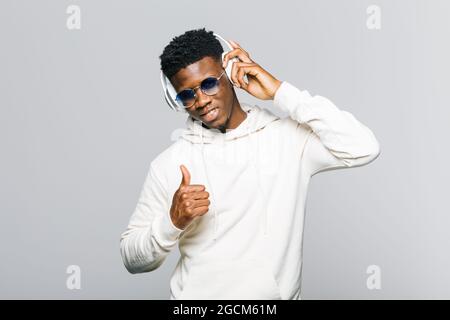 Happy young African American hipster guy in white hoodie and trendy sunglasses showing thumb up while listening to music through wireless headphones Stock Photo