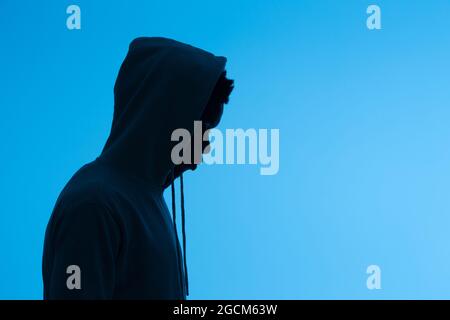 Side view of silhouette of unrecognizable African American male in hoodie standing on blue background in dark studio Stock Photo