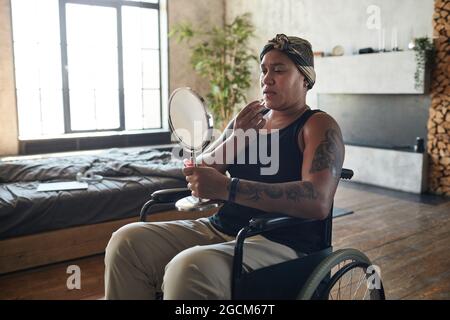 Portrait of African-American woman in wheelchair putting on makeup while holding mirror in home interior Stock Photo