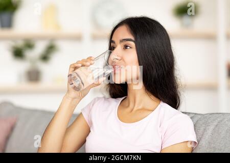 Keep hydrated concept. Attractive young Indian woman drinking water from glass on couch in living room Stock Photo