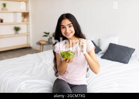 Weight loss, slimming diet concept. Happy Indian woman eating yummy vegetable salad on bed at home Stock Photo