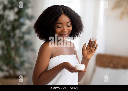 Young African American woman posing with jar of hydrating cream, smiling, wearing bath towel indoors Stock Photo