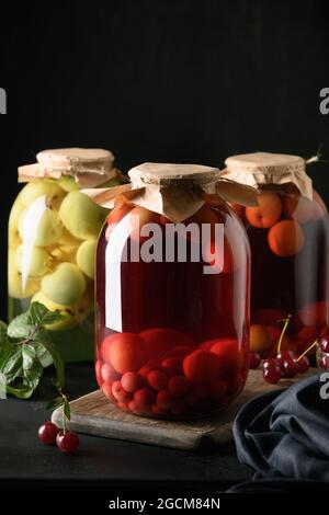 Three homemade canned cherry and apple compote in large glass jars on dark background. Vertical format. Stock Photo