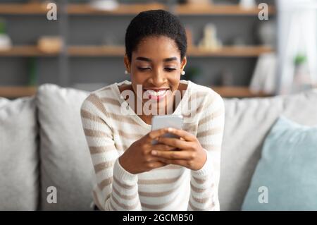 Cool App. Cheerful Young Black Woman Relaxing On Couch With Smartphone Stock Photo