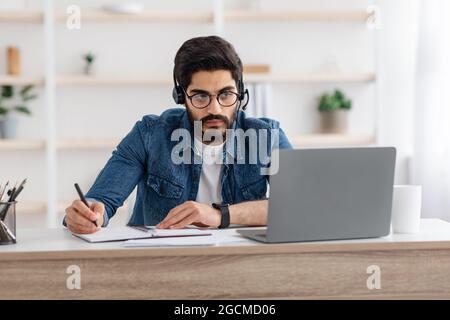 Millennial arab guy in headset watching online webinar on laptop at home, studying with computer and taking notes Stock Photo