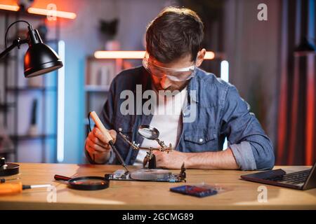 Focused bearded man in protective glasses sitting at table and soldering video card from modern laptop. Young male repairing important detail from computer by himself at home. Stock Photo