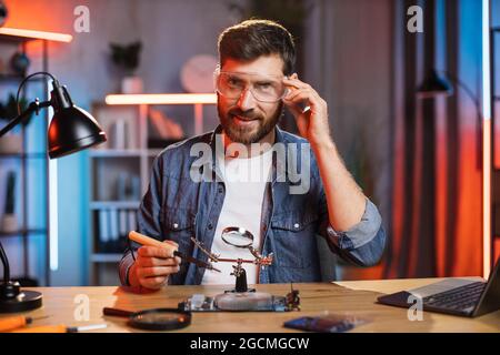 Caucasian young man in protective glasses using soldering iron and magnifying glass while repairing display card from modern laptop. Concept of people and technology. Stock Photo
