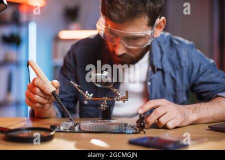 Concentrated repairman soldering non working video card from portable computer. Young bearded man in glasses fixing modern laptop at home. Stock Photo
