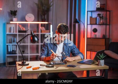 Handsome 35-aged bearded man in protective glasses sitting at table and soldering video card from modern laptop. Young male repairing important detail from computer by himself at home. Stock Photo