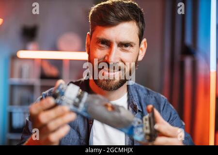 Close up of satisfied smiling bearded man sitting at table and showing repaired graphics card from modern laptop to camera. Young guy repairing device at home by himself Stock Photo