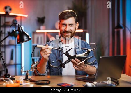 Smiling happy Caucasian bearded guy using soldering iron for repair modern quadcopter at home. Young man in casual clothes sitting at table and repairing drone. Stock Photo