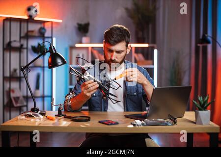 Concentrated bearded man using various tools for repairing modern quadcopter at home. Young caucasian guy in denim shirt fixing flying drone by himself. Stock Photo