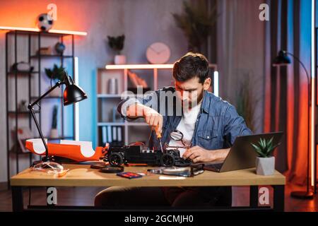 Bearded man using screwdriver and magnifying glass for fixing remote controlled toy car. Young guy sitting at table with various tools during repairing. Stock Photo