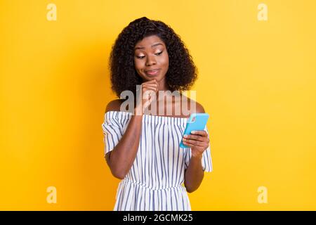 Photo of doubtful funny dark skin woman wear striped outfit arm chin looking modern gadget isolated yellow color background Stock Photo