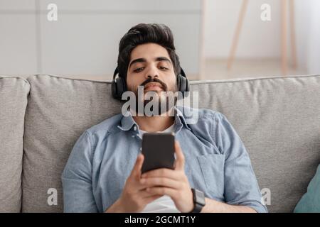 Listen to music with mobile app, watch videos, lesson and learn English online. Young arab guy in wireless headphones Stock Photo