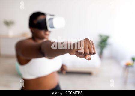 Virtual training. Overweight black lady in VR glasses using fighting simulator, making punch at home, focus on fist Stock Photo