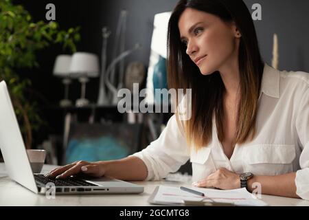 Beautiful Female Data Analyst sitting at the Table Works on a Laptop. Stylish Woman in the Modern Office Environment. Stock Photo