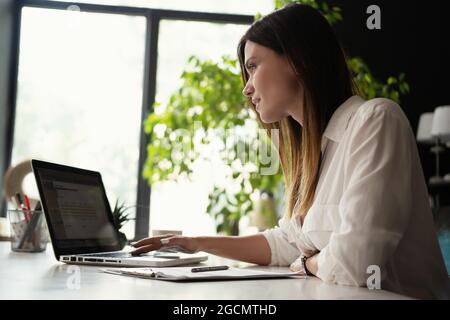 Beautiful Female Data Analyst sitting at the Table Works on a Laptop. Stylish Woman in the Modern Office Environment. Stock Photo