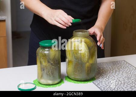 The woman closes the lid of a jar of cucumbers. Cucumbers are covered with pickling marinade. Harvest conservation. Stock Photo