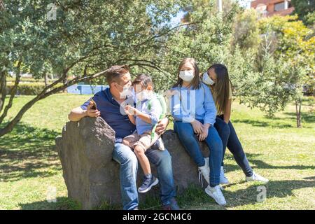 Family wearing mask sitting on a stone bench in a park, the father with the son on his legs and the mother with her daughter aside Stock Photo