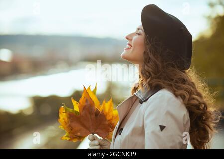 Hello november. relaxed trendy middle aged woman in beige trench coat with autumn yellow leaves outdoors in the city in autumn. Stock Photo