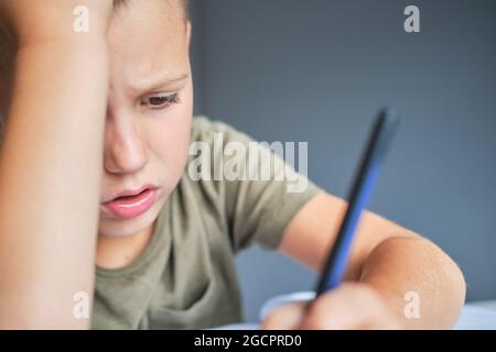 The bored and tired boy sitting at the table and using laptop while doing schoolwork at home. Homeschooling concept. Stock Photo