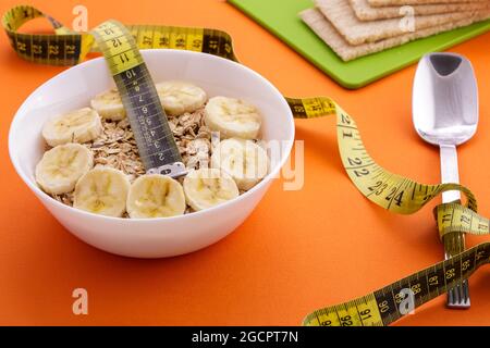 oatmeal with sliced banana and crispbread lie with spoon and yellow measuring tape on orange background, healthy food and diet concept Stock Photo