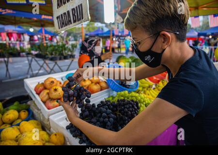 Young Malasian woman checks the quality of the grapes before buying. The girl is wearing face mask because of the virus crisis. Fresh or wet market in Stock Photo