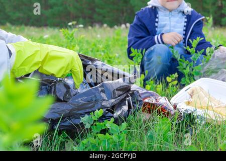 Female hands in yellow rubber gloves collect plastic trash in a black bag outdoors. In the background, a boy is helping. Selective focus. Summer sunny Stock Photo