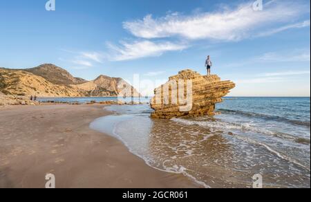 Young man standing on a rock, rocks in the water, sandy beach with rocky cliffs, Paralia Paradisos, Kos, Dodecanese, Greece Stock Photo