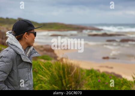 Malaysian woman looks over the pacific ocean at Anna Bay, Australia. Young asian girl with black baseball cap and warm coat looks out over the stormy Stock Photo