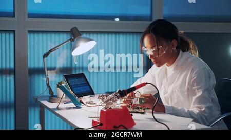 Close up of female electronics engineer in protective glasses checking motherboard with multimeter tester in advanced technology laboratory Stock Photo