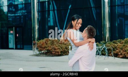 Happy boyfriend holding his black girlfriend in his hands and spinning around outdoors. An office building is in the background. Stock Photo