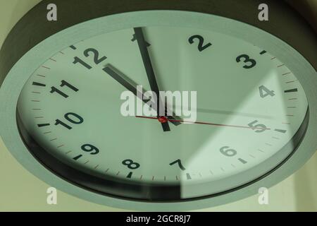 Modern white wall clock shows the time 5 after 12. Close up to a wall clock in sunlight with shadows on the  clock face. Time has expired. It is five Stock Photo