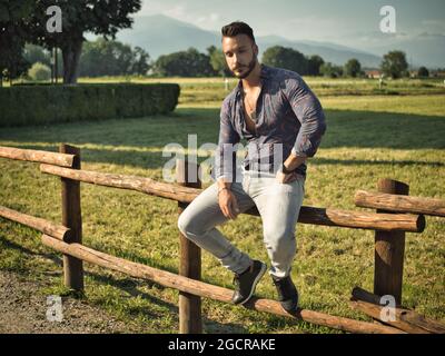 Stylish man sitting on fence in countryside Stock Photo