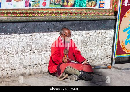 Lhasa, Tibet, China - November 15, 2019: Old Tibetan man  with  crutches and Tibetan Prayer Wheel sitting on the road side near the Jokhang Temple. El Stock Photo